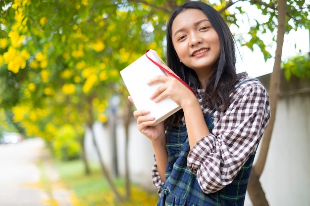 学生の若い女の子は美しい木の下で本を持っています。