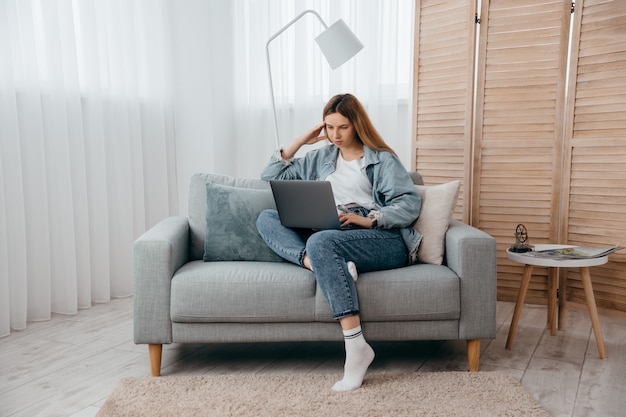 Student young girl lying on sofa at home and taking part in webinar getting new knowledge