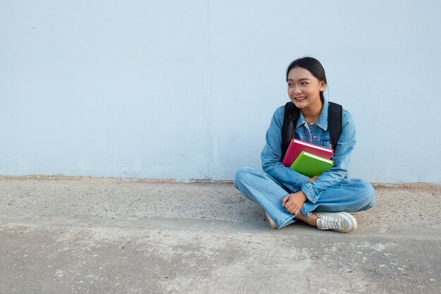 Student young girl sitting at school wear jean hold book with backpack blue background