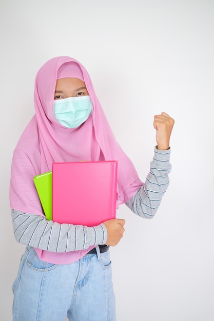 La giovane studentessa indossa l'hijab rosa e la maschera tiene i libri su sfondo bianco