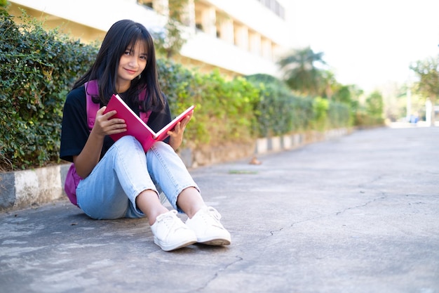 学校の背景を持つ床に座っているピンクのバックパックを持つ学生の若い女の子