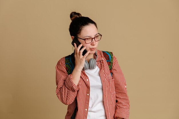 Student young woman in casual clothes wearing glasses with headphones and backpack talking on mobile phone with skeptic expression standing over brown background