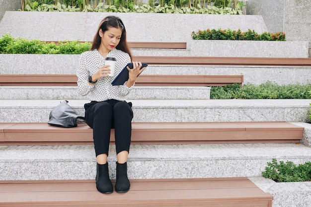 Student zittend op een bankje op de campus met een kopje koffie om mee te nemen en het lezen van e-book