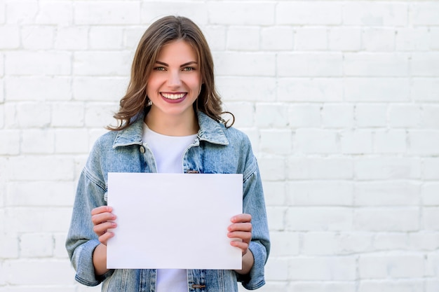 Studente die en een wit blad van document op een achtergrond van een witte bakstenen muur glimlacht houdt