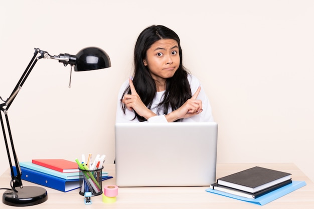 Studenten Aziatisch meisje in een werkplaats met laptop die op beige achtergrond wordt geïsoleerd die aan de lateralen richten die twijfels hebben