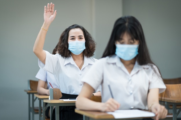 Studenten dragen gezichtsmaskerstudie in de klas en steken de hand op om de leraar te vragen tijdens de pandemie van het coronavirus. selectieve focus portret van universitaire studenten studeren in de klas.