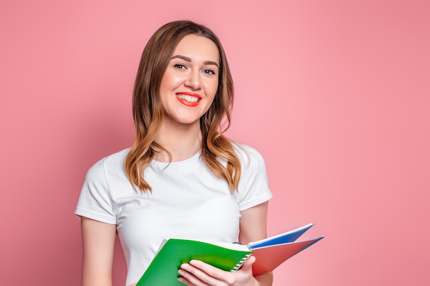 Studentenmeisje die een oefenboek, een notitieboekje en een glimlach houden die over roze achtergrond wordt geïsoleerd