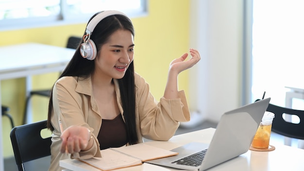 Studentenmeisje met een koptelefoon die online leert en een videogesprek voert via een laptopcomputer die in de coffeeshop zit.