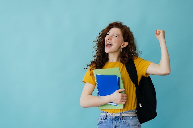 Studentenvrouw die op blauwe achtergrond wordt geïsoleerd die overwinningsgebaar toont