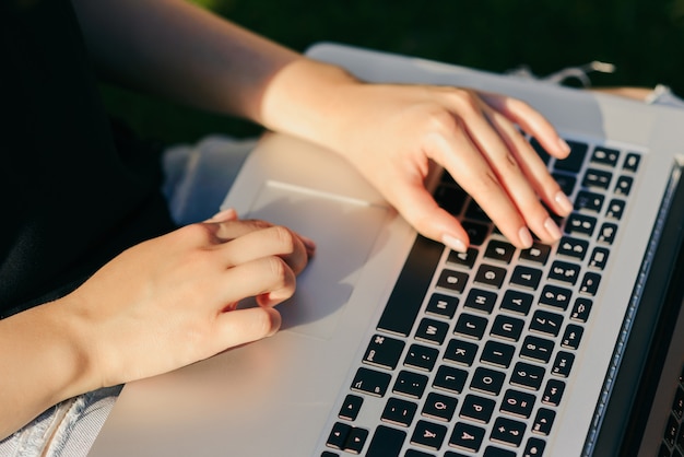 Studeren concept. Close-upmening die van vrouwelijke student in vrijetijdskleding op laptop `s toetsenbord typen, zittend op gras, in openlucht.