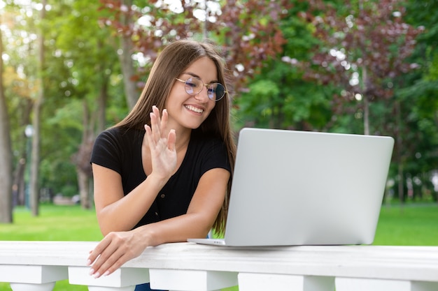 Studeren, frisse lucht, technologieconcept - Glimlachend jong oosters ogend meisje met een bril en vrijetijdskleding zwaait 'hallo' op een laptop terwijl ze in de zomer online lesgeeft in het park.