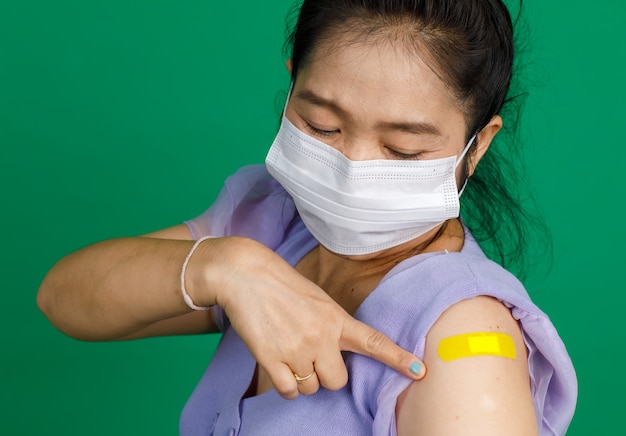 Studio close up shot of Asian female patient with face mask look at camera pointing at yellow bandage plaster after receive coronavirus covid 19 vaccination from doctor at clinic on green background.