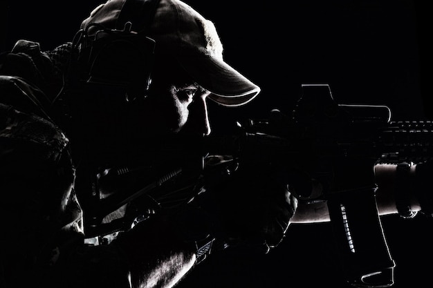 Studio contour achtergrondverlichting shot van special forces soldaat in uniformen en baseballcap, wijzend geweer, close-up portret op zwarte achtergrond