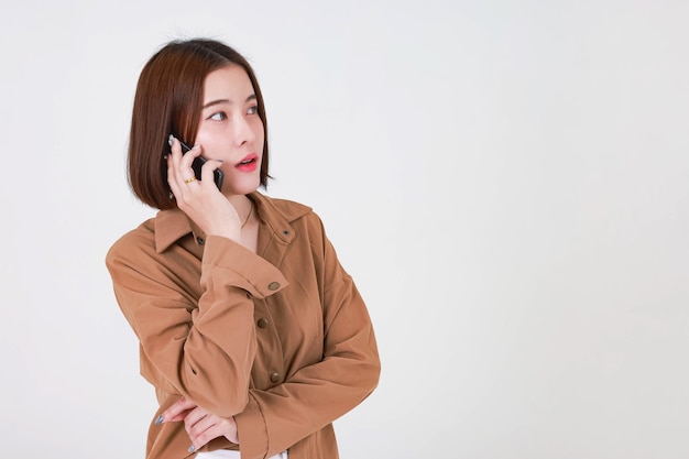 Studio ritagliato colpo di modello femminile asiatico giovane capelli piuttosto corti in camicia marrone a maniche lunghe in piedi sorridente tenendo lo smartphone su chiamata parlando parlando discutendo in chat su sfondo bianco.