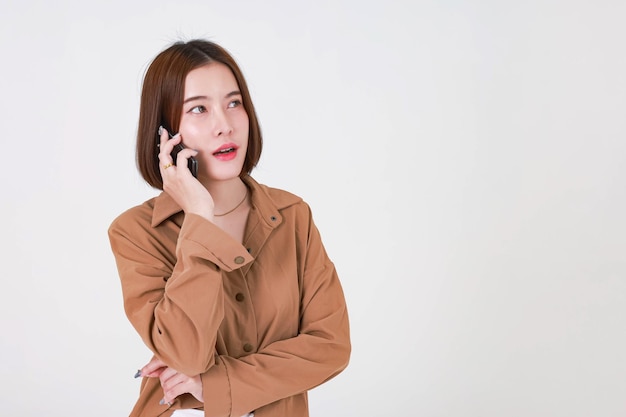 Studio cut out shot of Asian young pretty short hair female model in long sleeve brown shirt standing smiling holding smartphone on call talking speaking discussing chatting on white background.