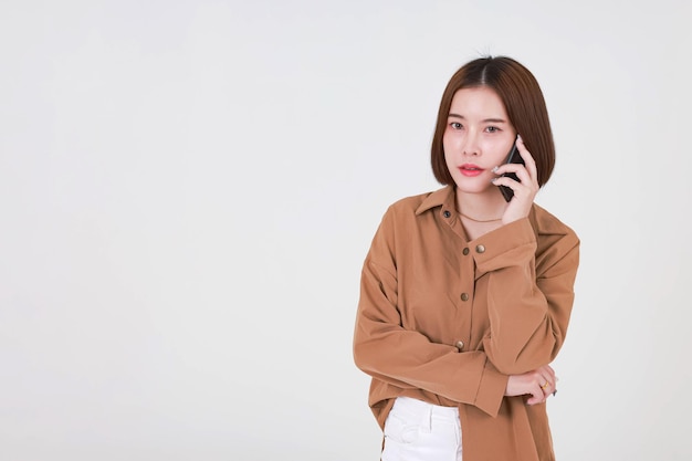 Studio cut out shot of Asian young pretty short hair female model in long sleeve brown shirt standing smiling holding smartphone on call talking speaking discussing chatting on white background.