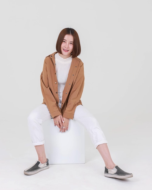 Studio cutout shot of Asian thoughtful young pretty short hair female model in long brown coat jacket casual shoes sitting crossed legs thinking decide solve idea on box chair on white background.