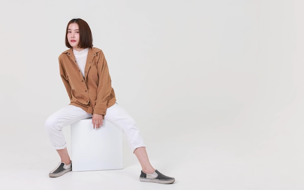 Studio cutout shot of Asian thoughtful young pretty short hair female model in long brown coat jacket casual shoes sitting crossed legs thinking decide solve idea on box chair on white background.