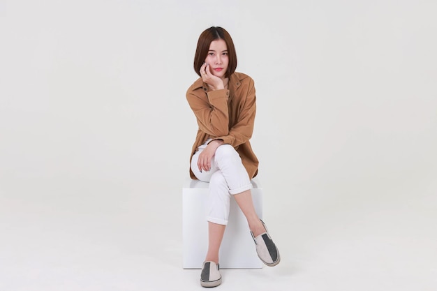 Studio cutout shot of Asian thoughtful young pretty short hair female model in long brown coat jacket casual shoes sitting crossed legs thinking decide solve idea on box chair on white background.