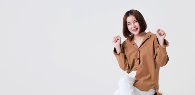 Studio cutout shot of Asian thoughtful young pretty short hair female model in long brown coat jacket casual shoes sitting crossed legs thinking decide solve idea on box chair on white background.