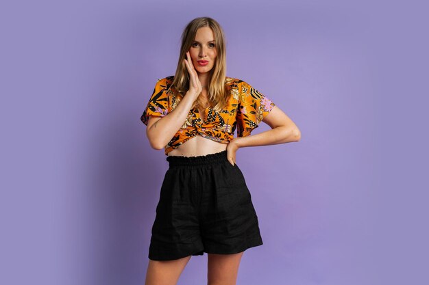 Studio fashion photo of pretty blond woman in stylish top and linen shorts posing on purple background