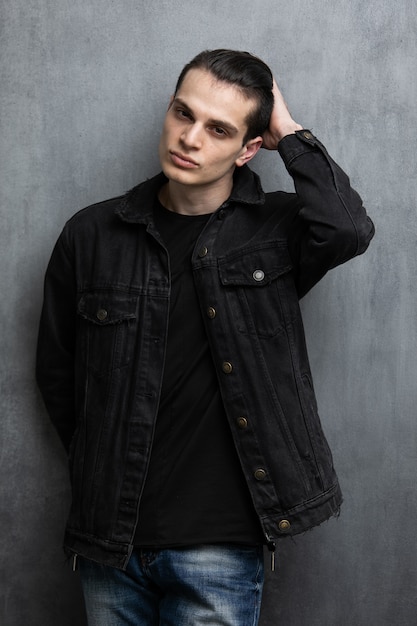 Studio fashion portrait of attractive young man in black jacket and blue jeans standing over grey textured wall.