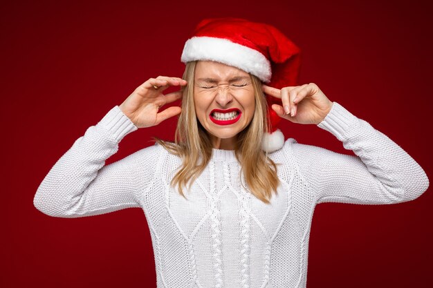 Studio foto van beklemtoonde jonge dame die haar ogen en oren sluit en ineenkrimpt terwijl ze de kerstmuts draagt