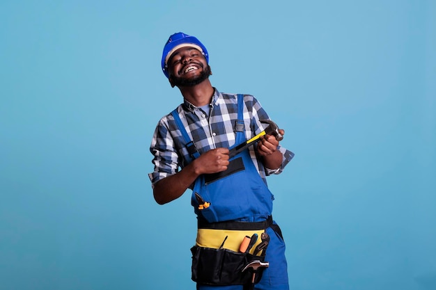 Studio foto van blije Afro-Amerikaanse bouwer in helm en gereedschapsriem met hamer met twee handen. Gelukkige bouwvakker die trots op blauwe achtergrond in studioschot glimlacht.