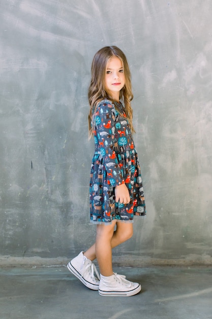 Studio image of lovely emotional caucasian teen girl in a blue dress and white sneakers on grey wall