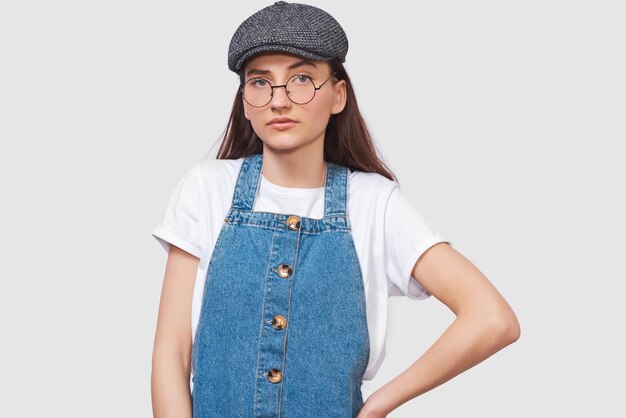 Studio image of serious young woman wears transparent trendy eyewear casual white tshirt denim dress trendy gray cap looks seriously directly into camera poses on white wall People emotions