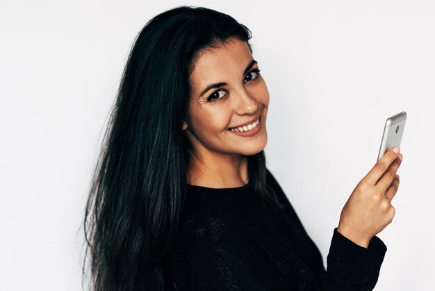 Studio image of smiling young woman types text messages on cell\
phone buying online via internet wears yellow sweater isolated on\
white studio wall technology and people concept