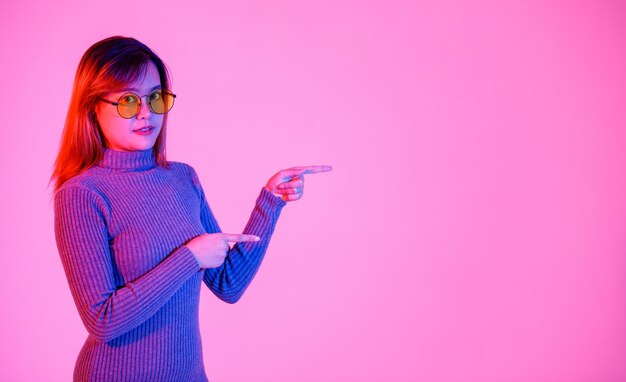 Studio-opname Aziatisch jong vrouwelijk model in grijze coltrui en oranje lenszonnebril die met wijsvingers naar lege kopieerruimte wijst en promotieproduct op roze lichte achtergrond presenteert.