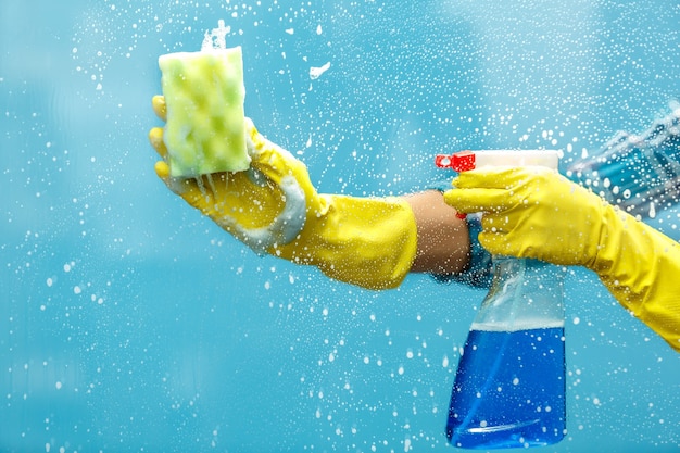 Studio opname door glas huishoudster. Vrouwenhand met handschoen die een fles spray vasthoudt en een spons gebruikt om het raam schoon te maken. Focus bij de hand