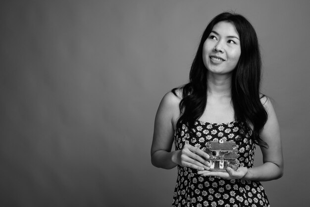 Studio-opname van een Aziatische vrouw die een jurk draagt terwijl ze een huisbeeldje vasthoudt tegen een grijze achtergrond in zwart-wit