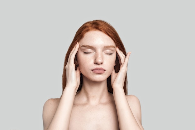 Studio-opname van een mooie roodharige vrouw die de ogen gesloten houdt en het hoofd met de handen aanraakt terwijl ze tegen een grijze achtergrond staat