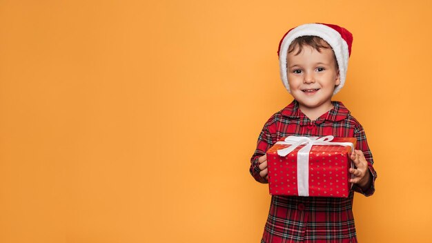 Studio photo of a baby boy in Christmas pajamas and a hat on a yellow background with a bright red gift box in his hands A place for your text advertising