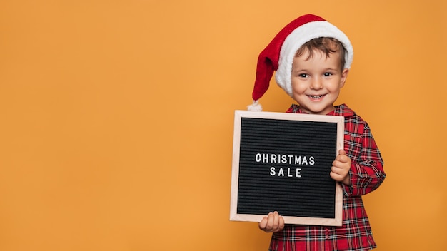 Studio photo of a baby boy in Christmas pajamas and a hat on a yellow background with a letter board with the text Merry Christmas in his hands. A place for your text, advertising.