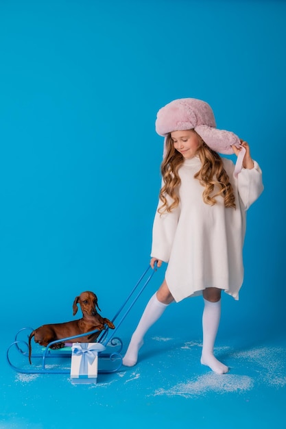 Studio photo of a girl in a white sweater and hat and a dachshund dog sitting in a sled