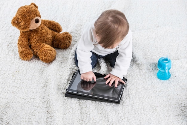 Studio photo with a white background of a baby touching the screen of a tablet with a teddy bear on the side