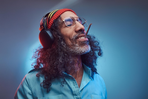 Studio portrait of African Rastafarian male enjoys music in headphones and smoking weed. Isolated on a blue background.