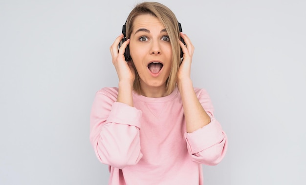 驚いた若い女性のスタジオポートレートは、白い背景で隔離のお気に入りの音楽を聞いている手でヘッドフォンを保持しているカメラを見てピンクのセーターを着ています広告用のスペースをコピーします