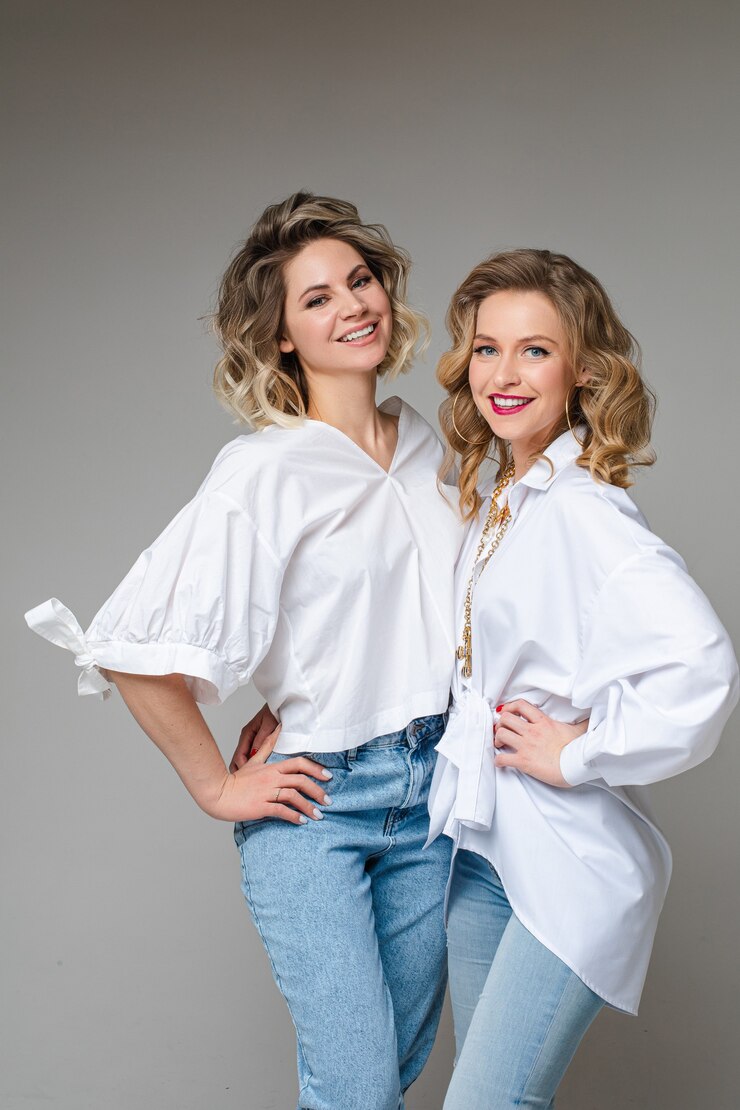 Photo studio portrait of attractive blonde adult women in white top and jeans posing snuggling on grey wall