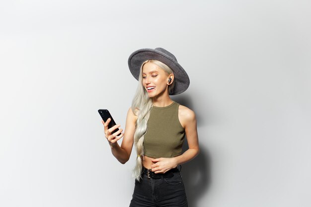 Studio portrait of beautiful happiness girl with wireless earphones into ears using smartphone holding device in hand wearing grey hat and shirt on white background