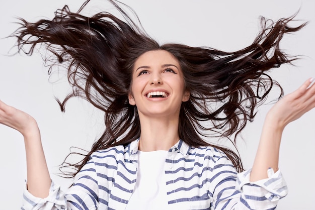 Studio portrait of beautiful joyful brunette woman with flying hair smiling and laughing looking up with raised hands posing over white background