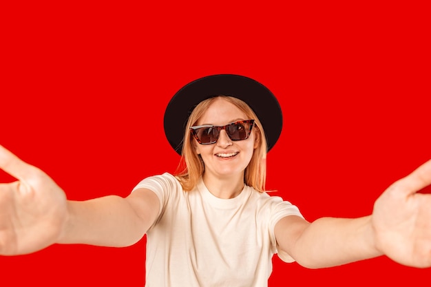 黒い帽子とサングラス白い歯に笑みを浮かべて、赤い背景の上に自分自身を撮影する selfie を作る美しい女性のスタジオ ポートレート