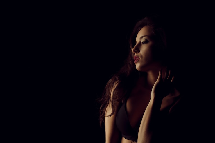 Photo studio portrait of beautiful young woman with bright makeup posing with naked shoulders in the dark