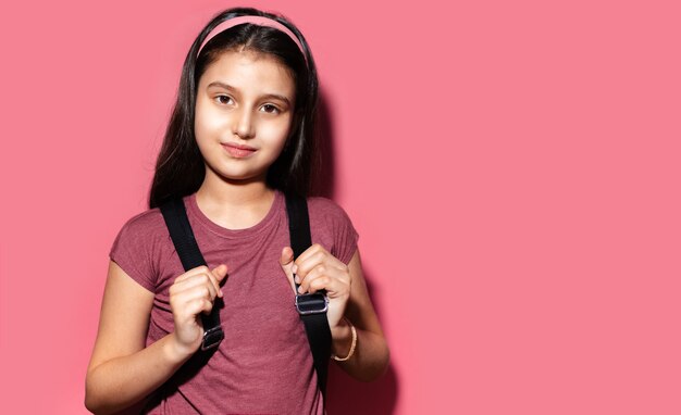 Ritratto dello studio della ragazza castana del bambino che si prepara per la scuola, zaino da portare e fascia su fondo di colore rosa pastello con lo spazio della copia.