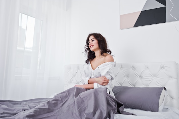 Studio portrait of brunette girl in white blouse lying on bed.