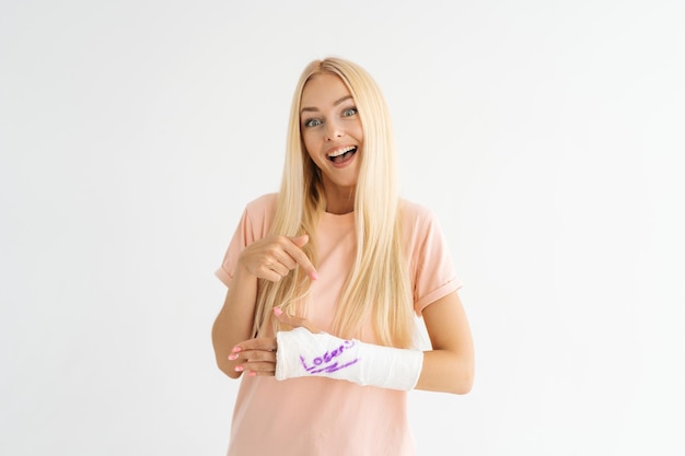 Studio portrait of cheerful injured young woman with broken arm wrapped in plaster bandage with funny inscription looking at camera on white background