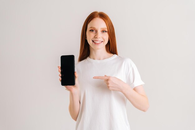 Studio portrait of cheerful redhead young woman with wide smile holding blank screen mobile phone and pointing index finger, looking at camera, standing on white isolated background.